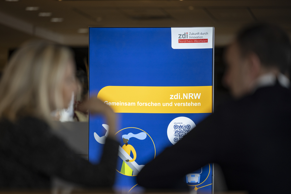 Zwei Personen sitzen vor einem blauen zdi.NRW-Schild, das den deutschen Slogan, einen QR-Code und Logos zeigt.