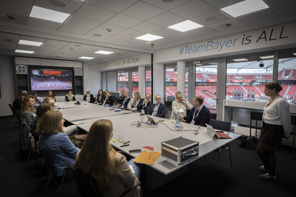 Menschen sitzen an einem Konferenztisch in einem modernen Besprechungsraum und hören einem Redner zu. Der Raum bietet einen Blick auf ein Sportstadion. An der Wand steht #TeamBayer.