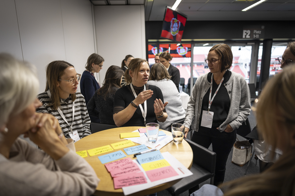 Mehrere Frauen sind um einen mit bunten Haftnotizen bedeckten Tisch versammelt und diskutieren in einem Veranstaltungsraum.