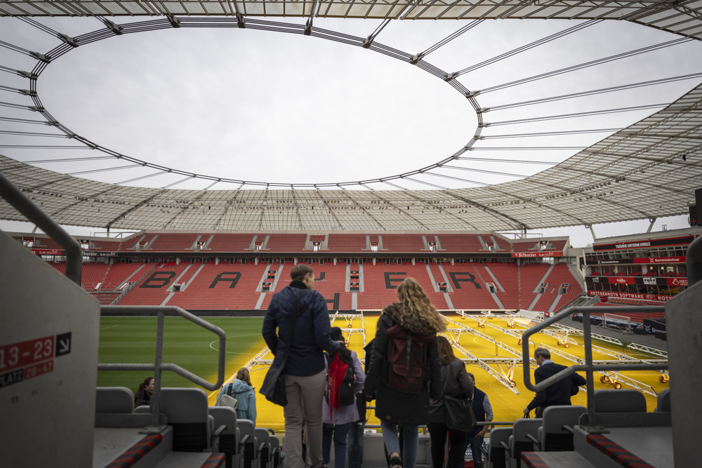 Die Menschen gehen die Stadiontreppe hinunter zum Spielfeld in einer leeren Arena mit einem teilweisen Dach und roten Sitzen mit der Aufschrift "BAYER".