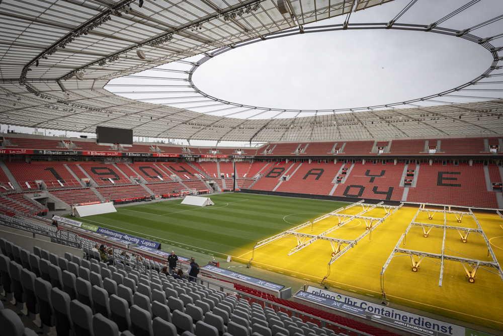 Das Stadion ist leer, mit roten Sitzen und "1904 BAYERN 04" auf der Tribüne. Künstliche Lichter erhellen Teile des grünen Feldes.