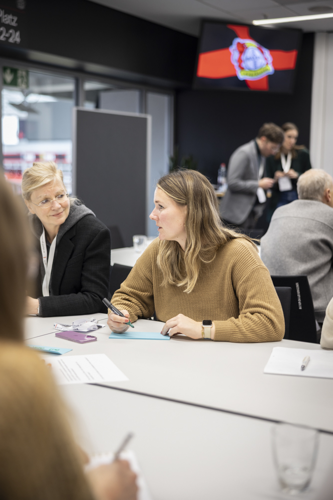 Zwei Frauen sitzen an einem Tisch mit Papieren und Stiften, während sie etwas besprechen. Andere Personen sind im Hintergrund zu sehen.