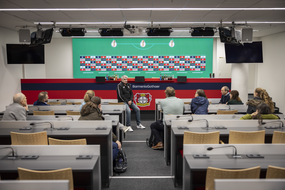 Menschen sitzen in einem Konferenzraum, vor einem Lautsprecher und vor einem Presse-Hintergrund mit dem Logo des Fußballvereins Bayer 04 Leverkusen.