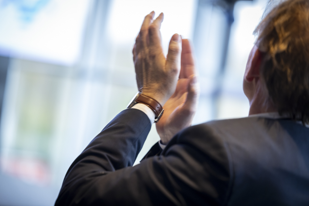 Eine Person im Anzug klatscht, von hinten gesehen. Im Hintergrund erscheinen unscharfe Fenster.