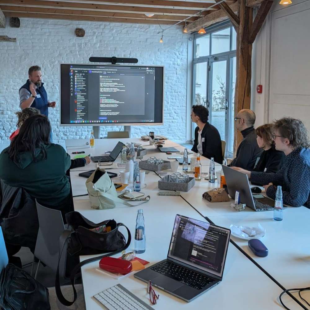 In einem modernen Besprechungsraum sitzen Menschen mit Laptops und Flaschen um einen Tisch herum, während eine Person etwas auf einem großen Bildschirm präsentiert.