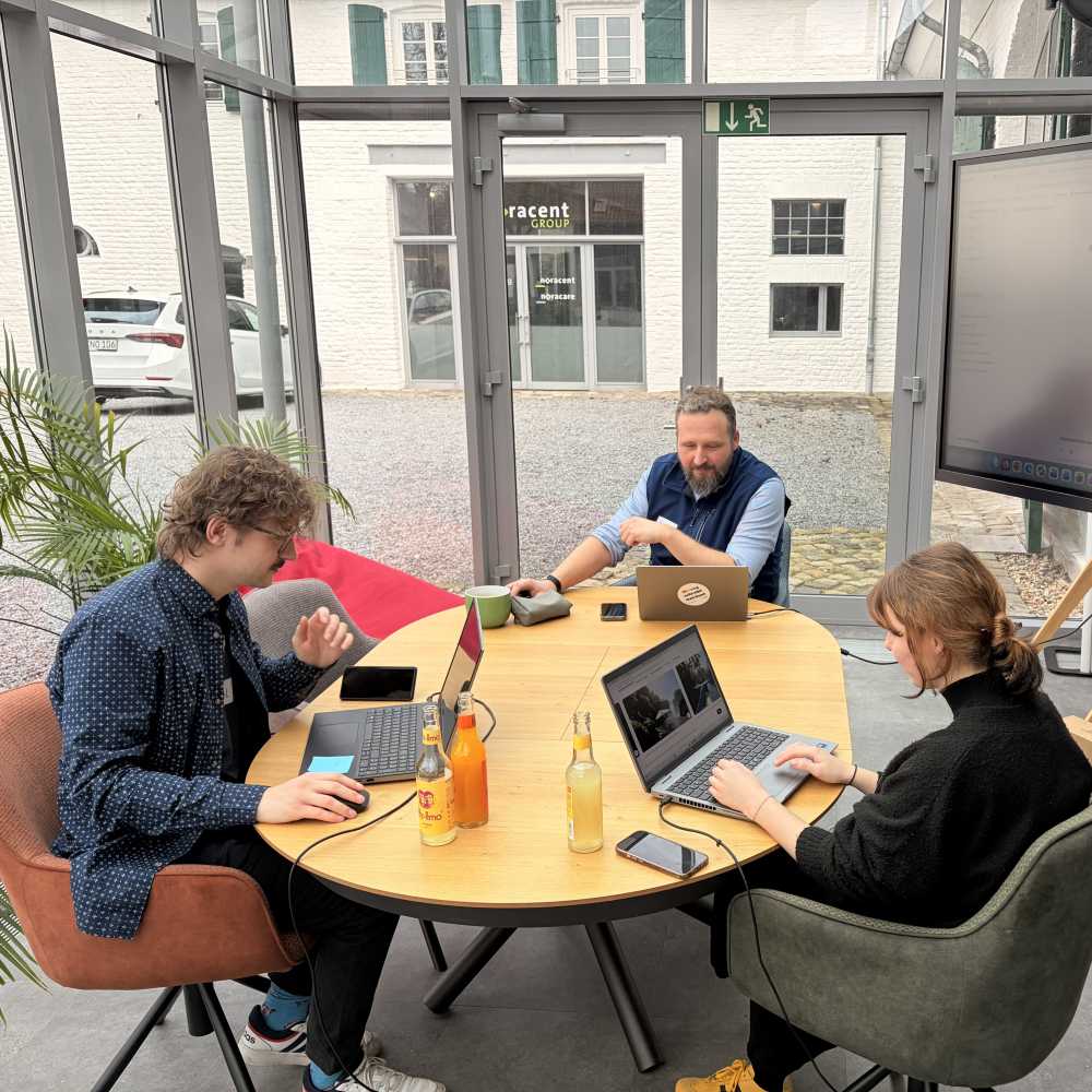 Drei Personen sitzen an einem runden Tisch und arbeiten mit Laptops. In einem hellen Raum mit großen Fenstern stehen Getränke auf dem Tisch.