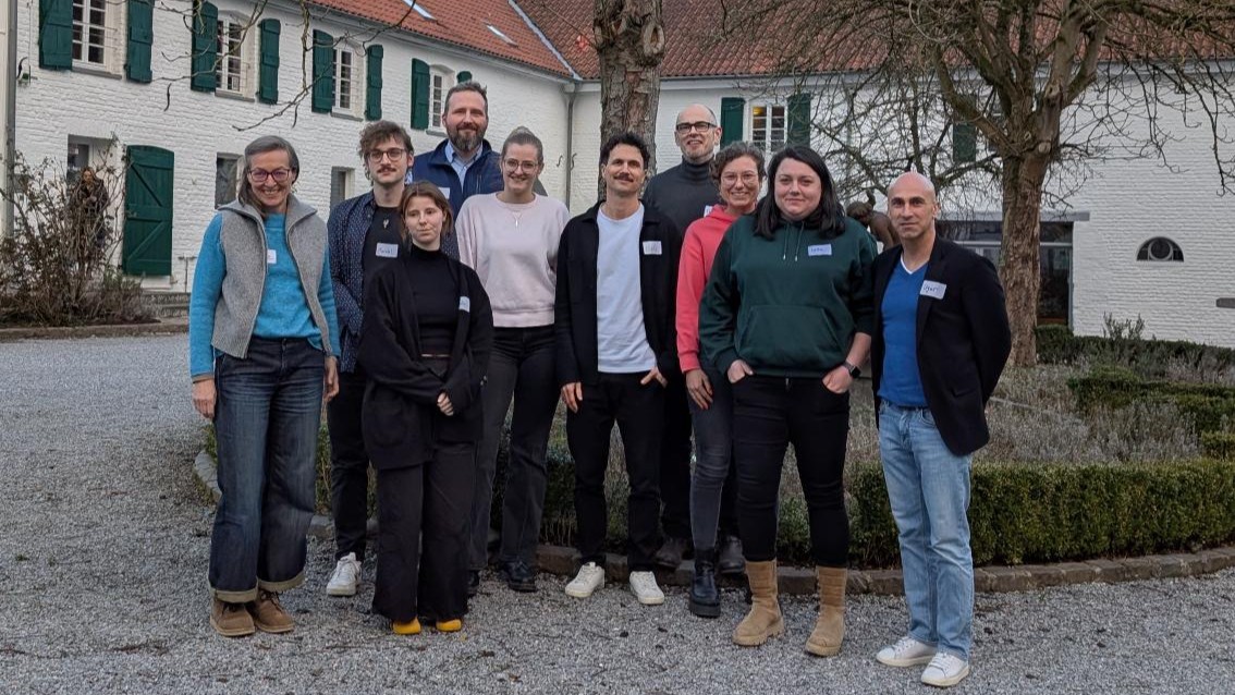Zehn Personen stehen draußen und posieren für ein Foto vor einer runden Hecke und einem großen weißen Gebäude mit grünen Fensterläden.