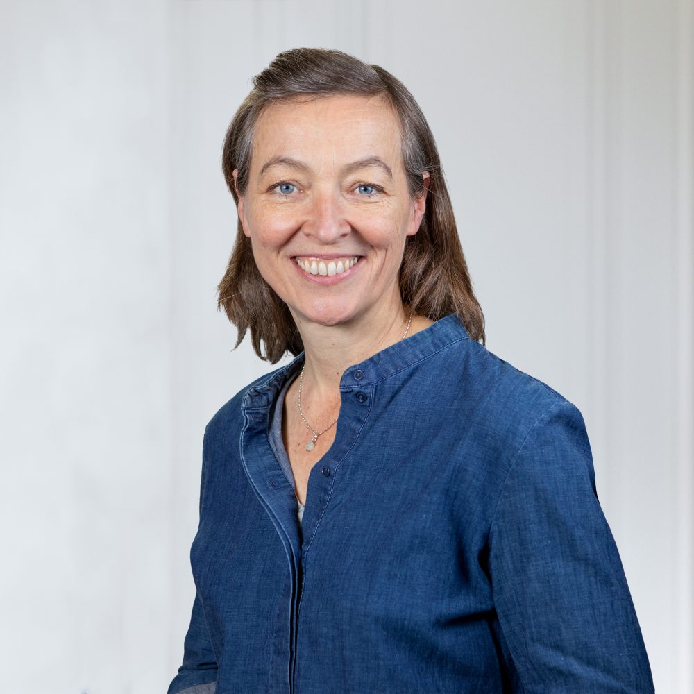 Eine Frau mit kurzen braunen Haaren lächelt. Sie trägt ein blaues Button-up-Hemd und steht vor einem schlichten hellen Hintergrund.