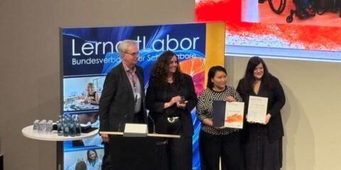 Vier Personen stehen auf einer Bühne und halten Urkunden in der Hand. In der Nähe steht ein Podium, und im Hintergrund ist ein "Lernort Labor"-Transparent zu sehen.
