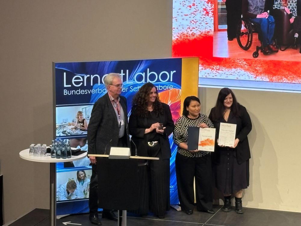 Vier Personen stehen auf einer Bühne und halten Urkunden in der Hand. In der Nähe steht ein Podium, und im Hintergrund ist ein "Lernort Labor"-Transparent zu sehen.