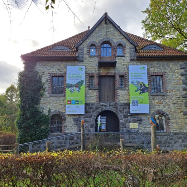 Das Bild zeigt ein Steingebäude mit roten Dachziegeln. An der Fassade hängen zwei große Banner, und eine Holzrampe führt zum Eingang. Das Gebäude ist von Bäumen umgeben.