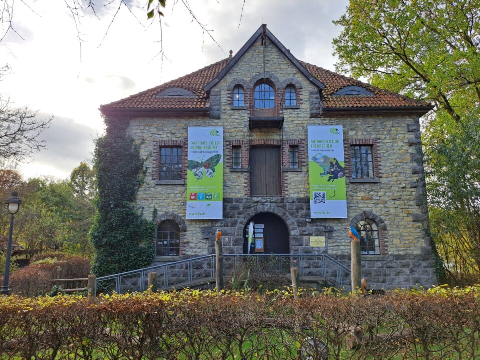 Das Bild zeigt ein Steingebäude mit roten Dachziegeln. An der Fassade hängen zwei große Banner, und eine Holzrampe führt zum Eingang. Das Gebäude ist von Bäumen umgeben.