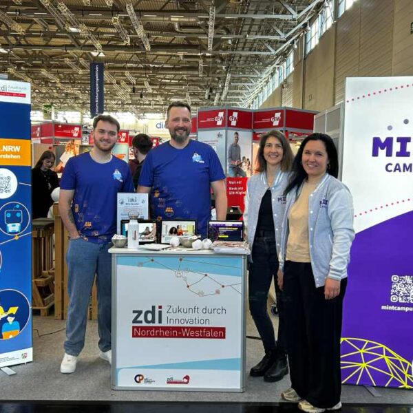 Vier Personen stehen an einem Stand von zdi.NRW und MINT-Campus auf einer Indoor-Veranstaltung. Um sie herum sind Banner und Materialien ausgelegt.