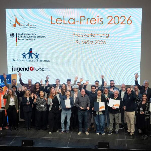 Bei der Verleihung des LeLa-Preises 2026 stehen mehrere Personen mit Urkunden in der Hand auf der Bühne. Hinter ihnen sind ein Dia und Banner zu sehen.