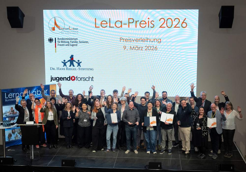 Bei der Verleihung des LeLa-Preises 2026 stehen mehrere Personen mit Urkunden in der Hand auf der Bühne. Hinter ihnen sind ein Dia und Banner zu sehen.