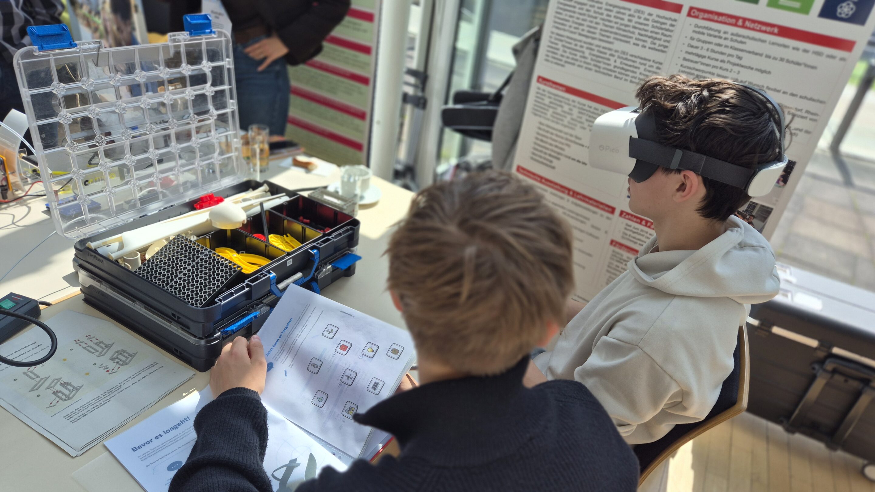 Zwei Jungen sitzen an einem Tisch mit technischen Geräten. Der eine trägt ein VR-Headset, der andere schaut in ein aufgeschlagenes Handbuch. Hinter ihnen stehen wissenschaftliche Poster.