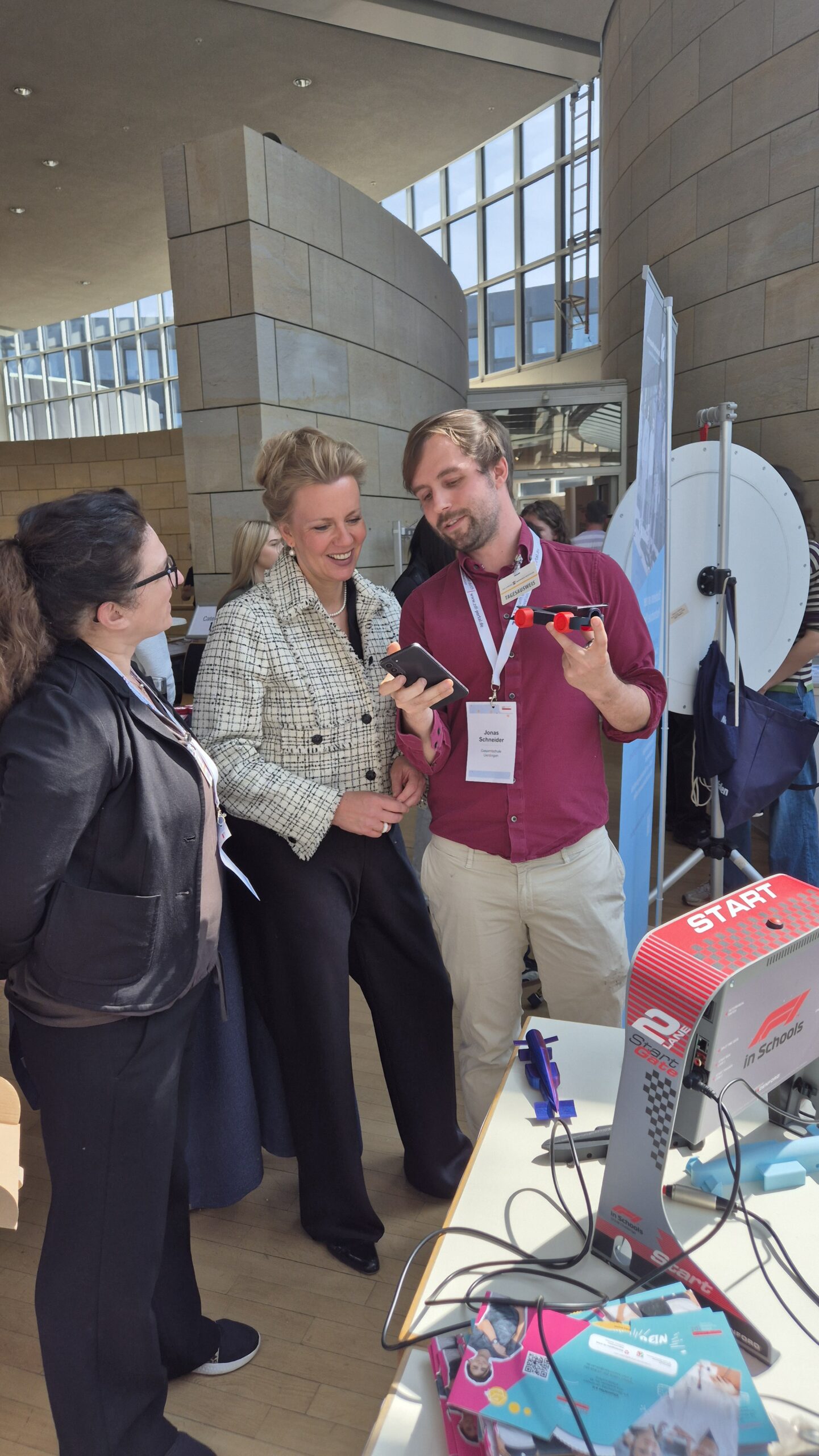 Drei Personen stehen in einem Innenraum an einem Ausstellungstisch. Sie schauen auf ein Telefon und ein 3D-gedrucktes Objekt. Veranstaltungsplaketten sind sichtbar.