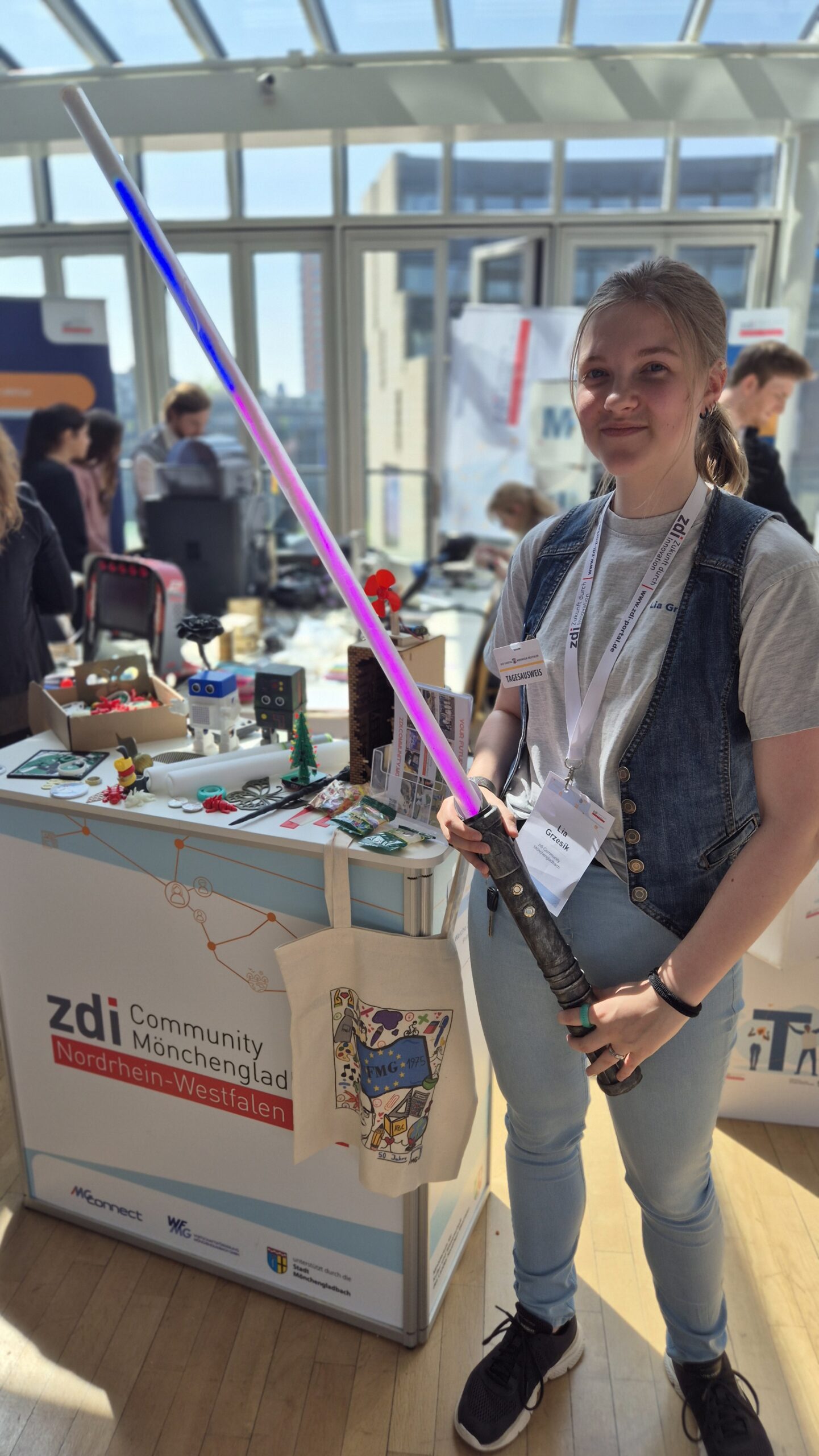 Ein Jugendlicher hält ein Spielzeug-Lichtschwert neben einem Display der zdi-Gemeinschaft Mönchengladbach bei einer Wissenschaftsveranstaltung in der Hand.