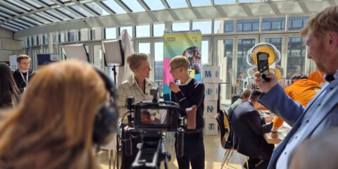 Bei einer Indoor-Veranstaltung interagieren Menschen und nehmen Videos auf. In einem hellen, glasüberdachten Raum sind Banner, eine Kamera und Beleuchtungsausrüstung aufgebaut.