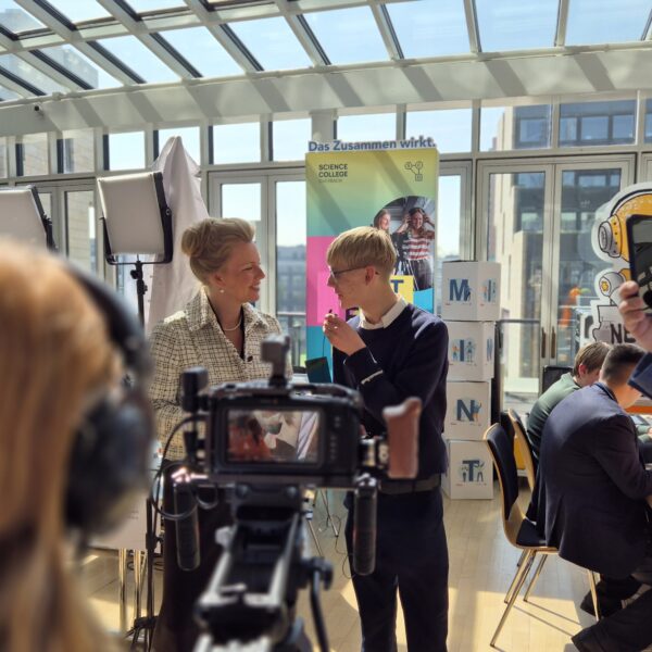 Bei einer Indoor-Veranstaltung interagieren Menschen und nehmen Videos auf. In einem hellen, glasüberdachten Raum sind Banner, eine Kamera und Beleuchtungsausrüstung aufgebaut.