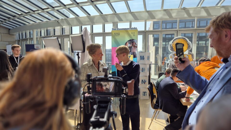 Bei einer Indoor-Veranstaltung interagieren Menschen und nehmen Videos auf. In einem hellen, glasüberdachten Raum sind Banner, eine Kamera und Beleuchtungsausrüstung aufgebaut.