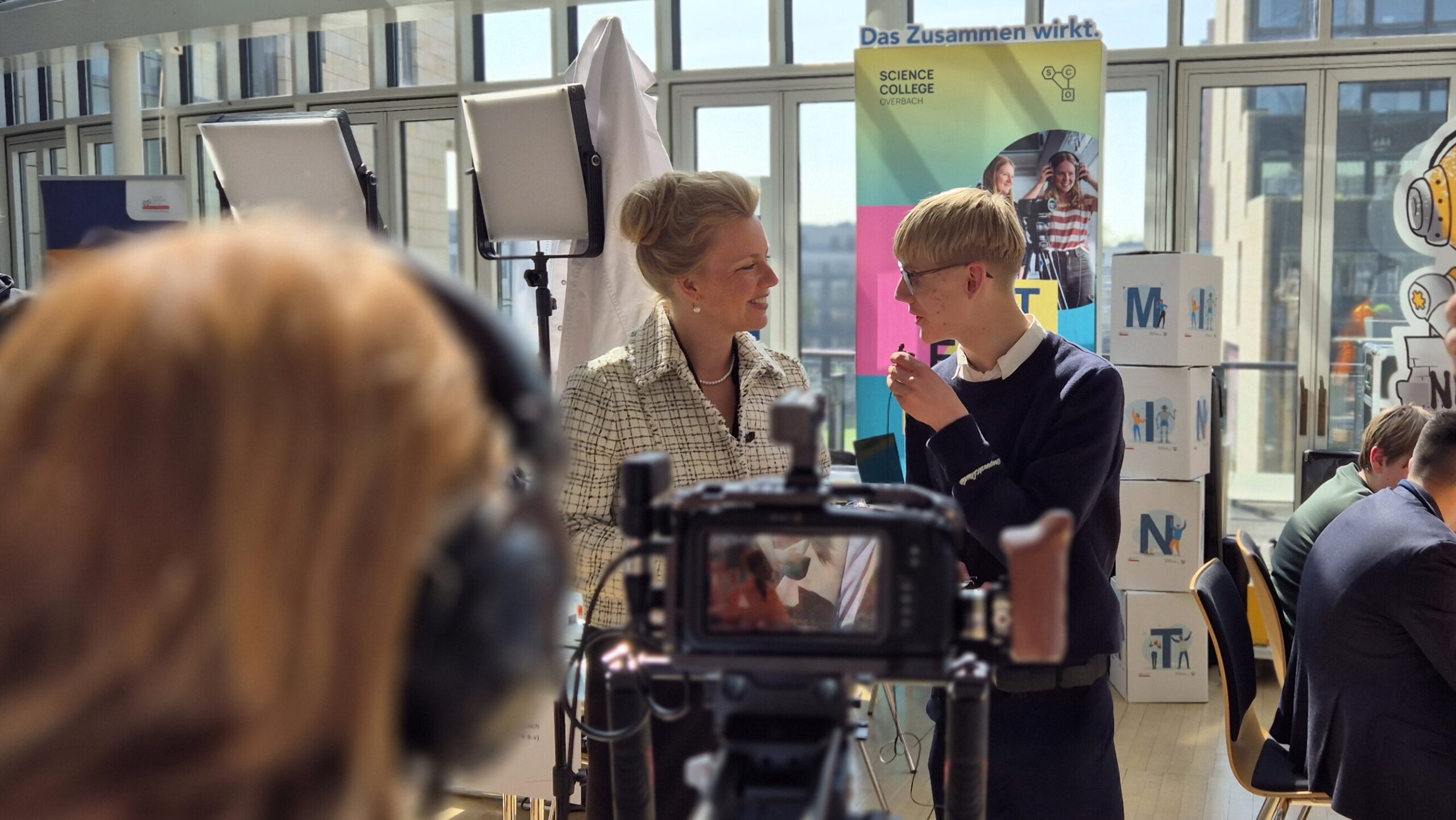 Bei einer Indoor-Veranstaltung interagieren Menschen und nehmen Videos auf. In einem hellen, glasüberdachten Raum sind Banner, eine Kamera und Beleuchtungsausrüstung aufgebaut.