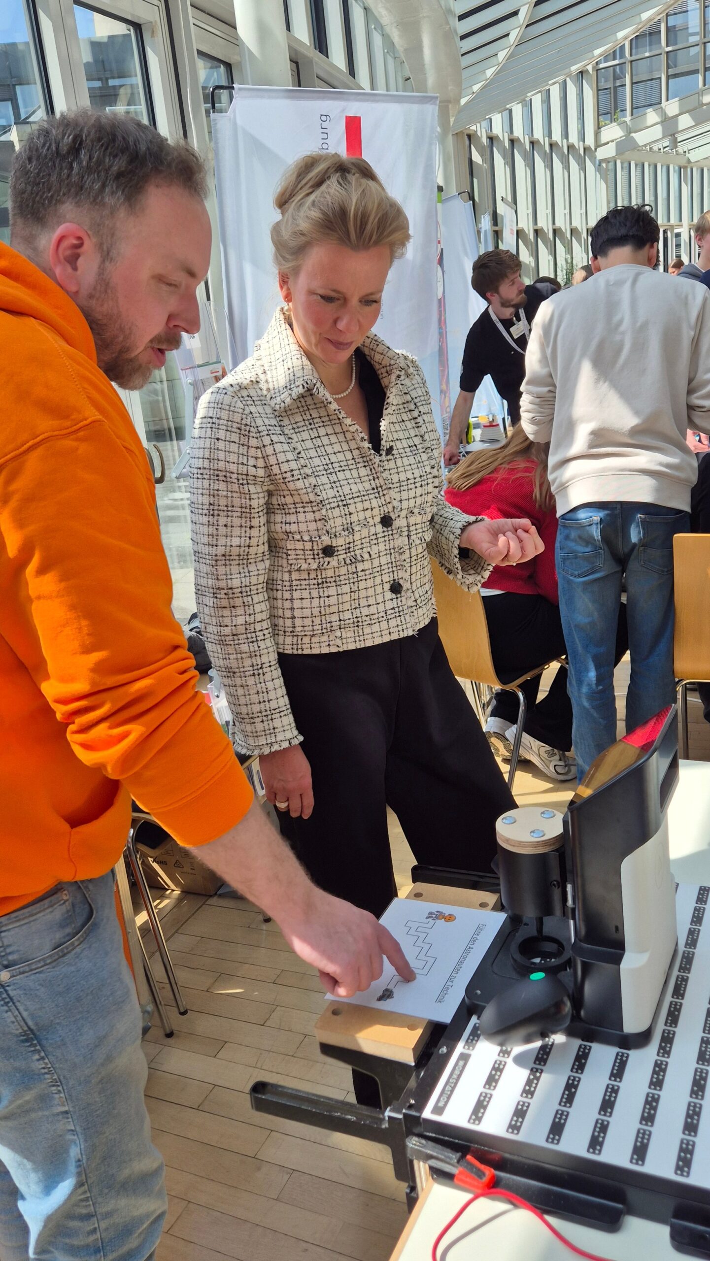 Ein Mann demonstriert einer Frau bei einer Indoor-Veranstaltung eine Maschine und zeigt ihr ein bedrucktes Blatt. Im Hintergrund sind Menschen und Tische zu sehen.