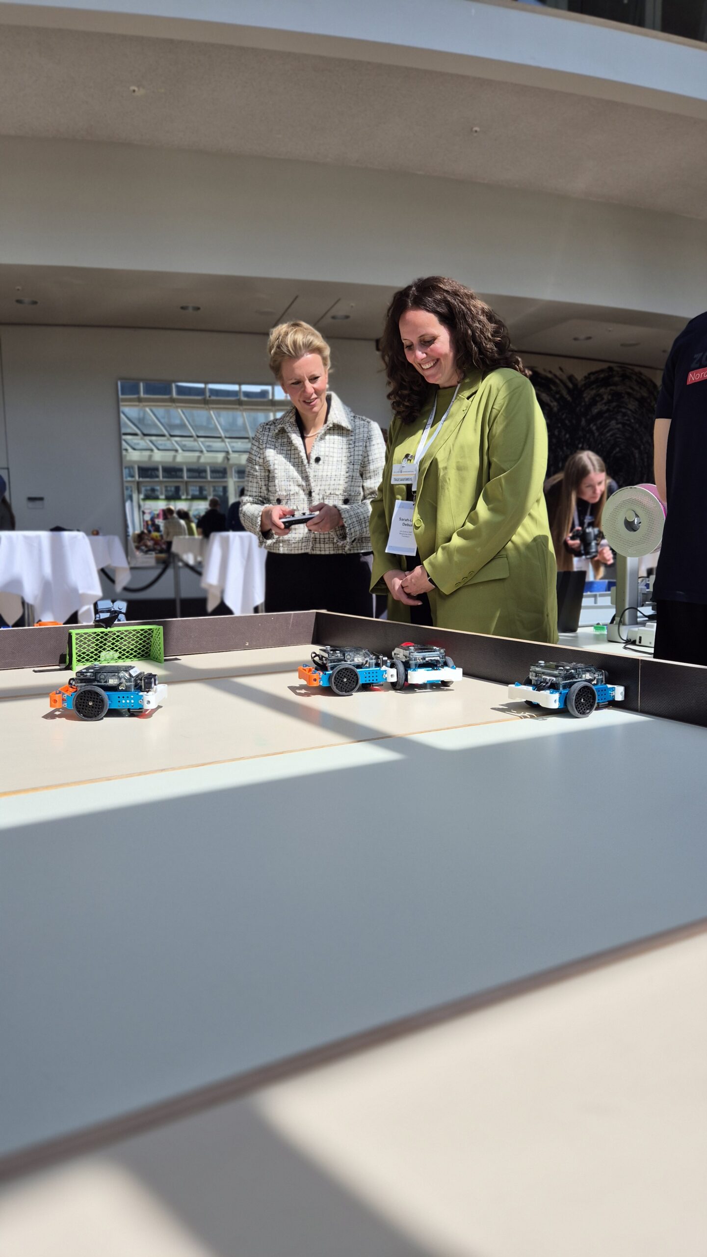 Zwei Frauen beobachten kleine Roboterfahrzeuge auf einem Tisch in einem hell erleuchteten Raum, hinter dem andere Menschen und Tische zu sehen sind.