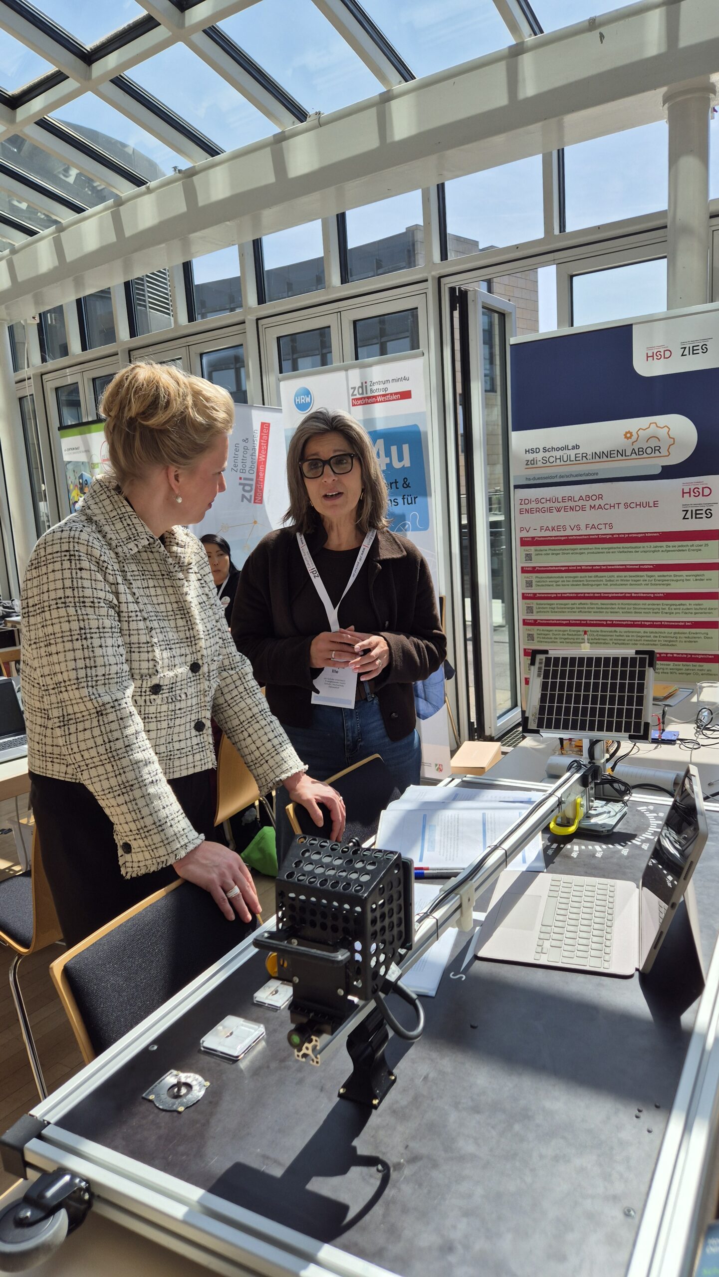 Zwei Frauen unterhalten sich bei einer Indoor-Veranstaltung an einem Tisch mit technischen Geräten und einem Laptop, hinter denen Informationsdisplays angebracht sind.