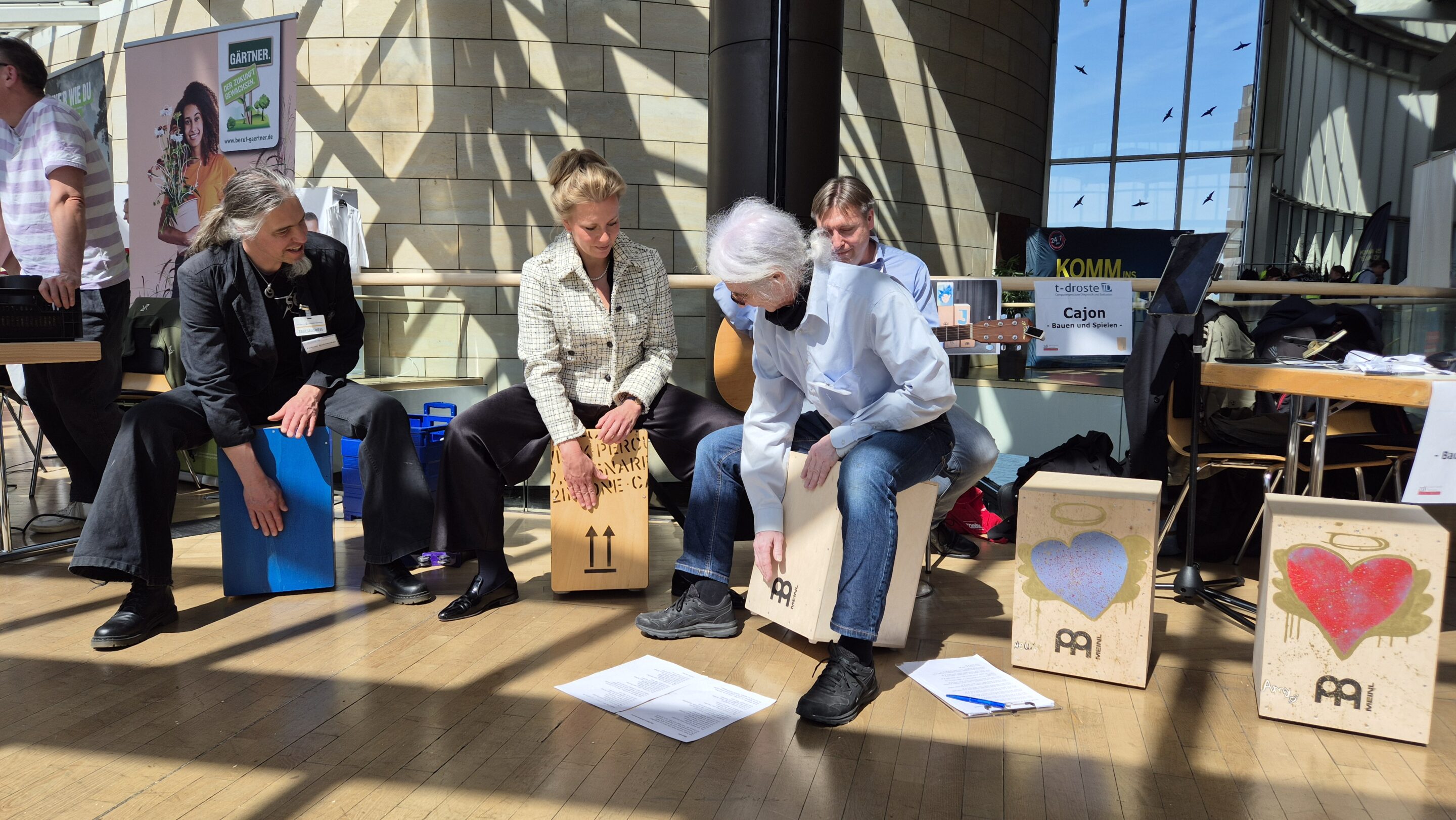 Vier Personen sitzen auf Cajón-Trommeln in einem Raum und studieren Papiere auf dem Boden. Im sonnenbeschienenen Hintergrund sind bemalte Cajóns und Fahnen zu sehen.