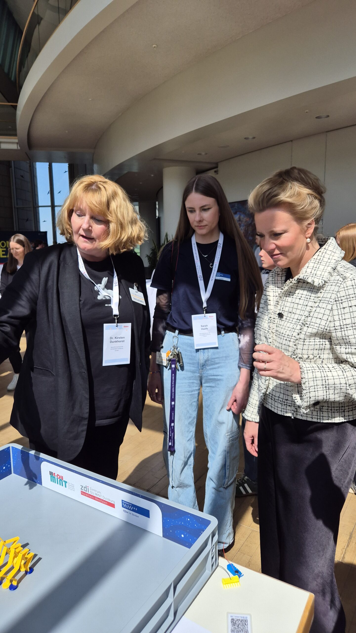 Drei Frauen mit Namensschildern stehen bei einer Indoor-Veranstaltung um einen Tisch mit Lehrmaterial, während das Sonnenlicht durch große Fenster strömt.