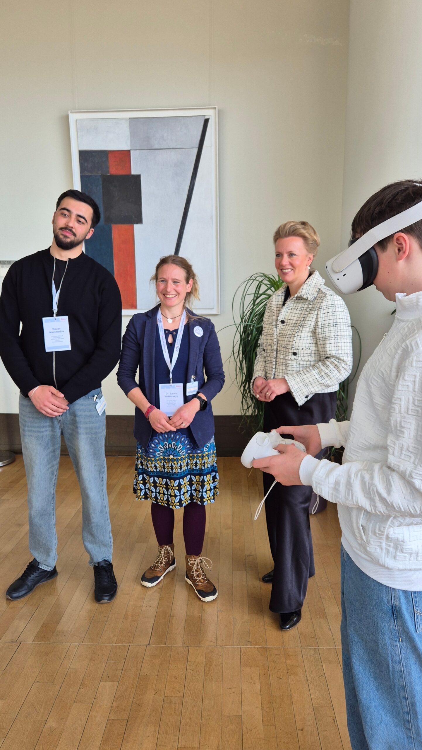 Vier Personen stehen in einem Raum. Drei lächeln und posieren für ein Foto, während die vierte Person auf der rechten Seite ein VR-Headset und einen Controller benutzt.