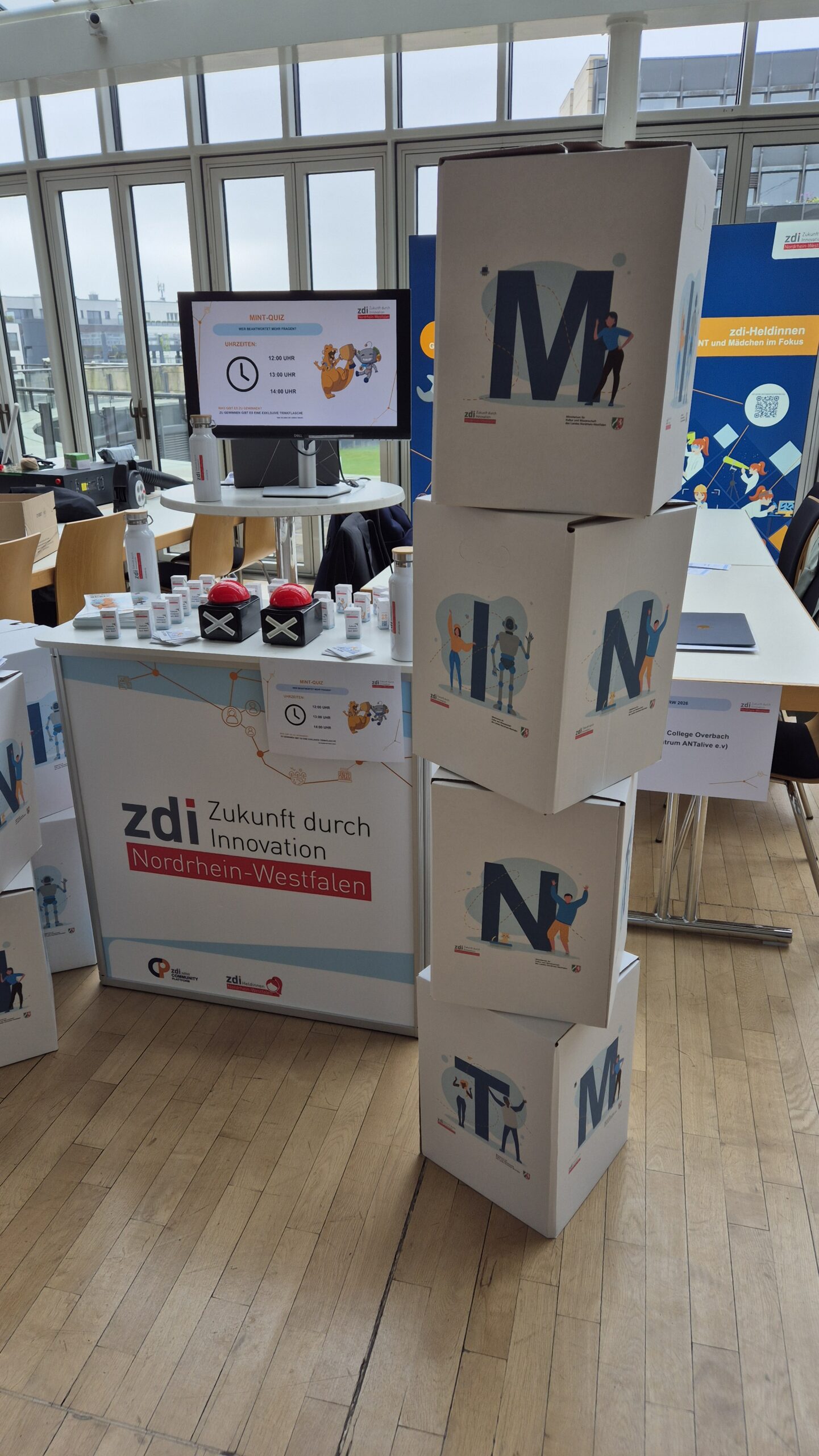 Das Bild zeigt einen Messestand mit gestapelten Kartons, einem Tisch mit Werbeartikeln und einem Bildschirm für "zdi Zukunft durch Innovation NRW".