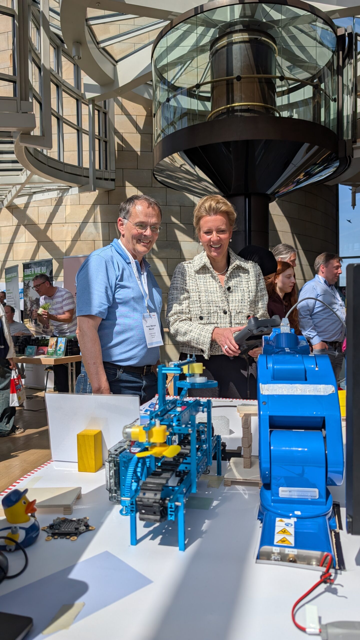 Zwei Personen stehen bei einer Indoor-Veranstaltung neben einem Ausstellungstisch mit blauen und gelben Robotermaschinen. Im Hintergrund sind andere Teilnehmer zu sehen.