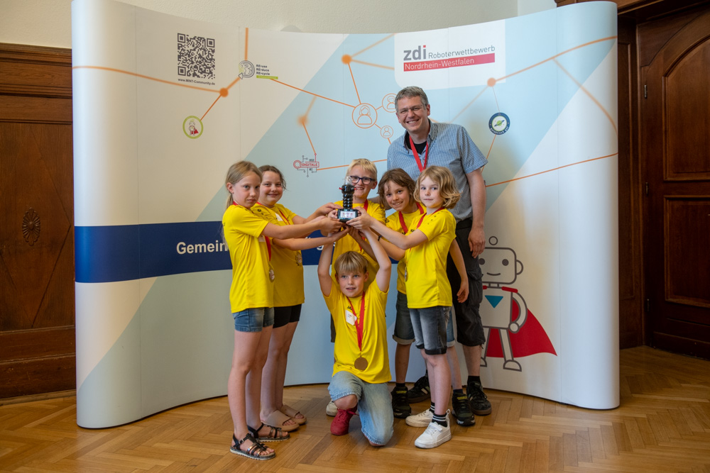 Die Kinder in gelben T-Shirts und ein Mann stehen in einem Raum, halten eine kleine Trophäe in den Händen und lächeln vor einem Hintergrund mit MINT-Motiven.