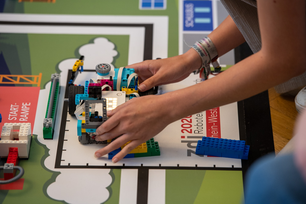Eine Person baut auf einer Matte, auf der „zdi 2023 Roboter Rhein-Westfalen“ steht, einen LEGO-Roboter zusammen.