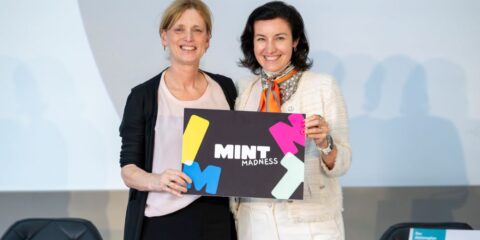 Zwei Frauen lächeln nebeneinander und halten vor einem schlichten Hintergrund ein Schild mit der Aufschrift „MINT MADNESS“ in die Kamera, um den Aktionsplan MINT+ zu feiern.