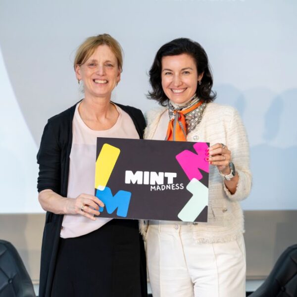 Zwei Frauen lächeln nebeneinander und halten vor einem schlichten Hintergrund ein Schild mit der Aufschrift „MINT MADNESS“ in die Kamera, um den Aktionsplan MINT+ zu feiern.