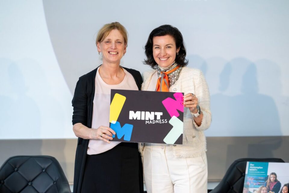 Zwei Frauen lächeln nebeneinander und halten vor einem schlichten Hintergrund ein Schild mit der Aufschrift „MINT MADNESS“ in die Kamera, um den Aktionsplan MINT+ zu feiern.