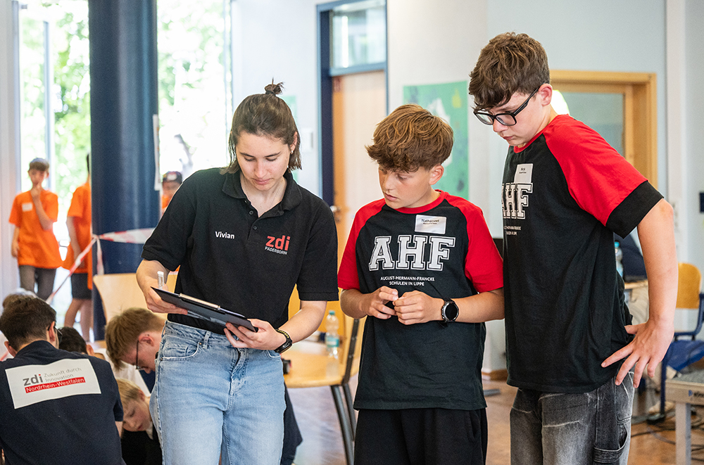 Eine Frau zeigt zwei Jungen in identischen T-Shirts bei einer Veranstaltung in der Halle ein Tablet. Im Hintergrund unterhalten sich andere Personen miteinander.
