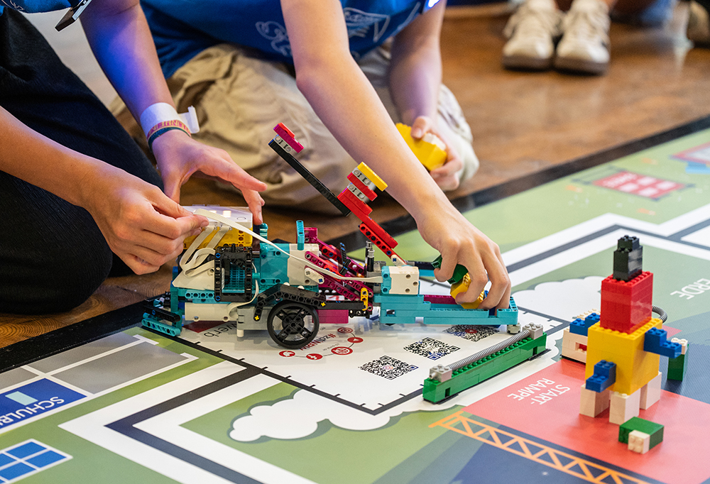 Zwei Personen bauen einen kleinen LEGO-Roboter auf einer Wettkampfmatte zusammen und nehmen ihn in Betrieb. Daneben steht eine Konstruktion aus Bausteinen.