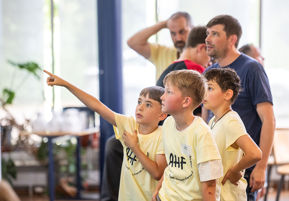 Drei Jungen in einheitlichen gelben T-Shirts stehen beieinander, einer zeigt nach vorne. Im hellen Hintergrund in einem Innenraum sind zwei Erwachsene und ein Kind zu sehen.