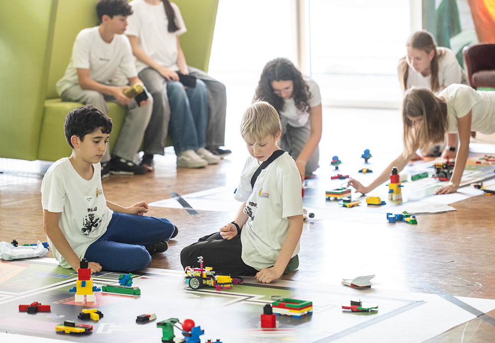 Kinder in weißen T-Shirts sitzen und knien auf dem Boden. Sie bauen mit bunten LEGO-Steinen und arbeiten in einem hellen Raum an Projekten.