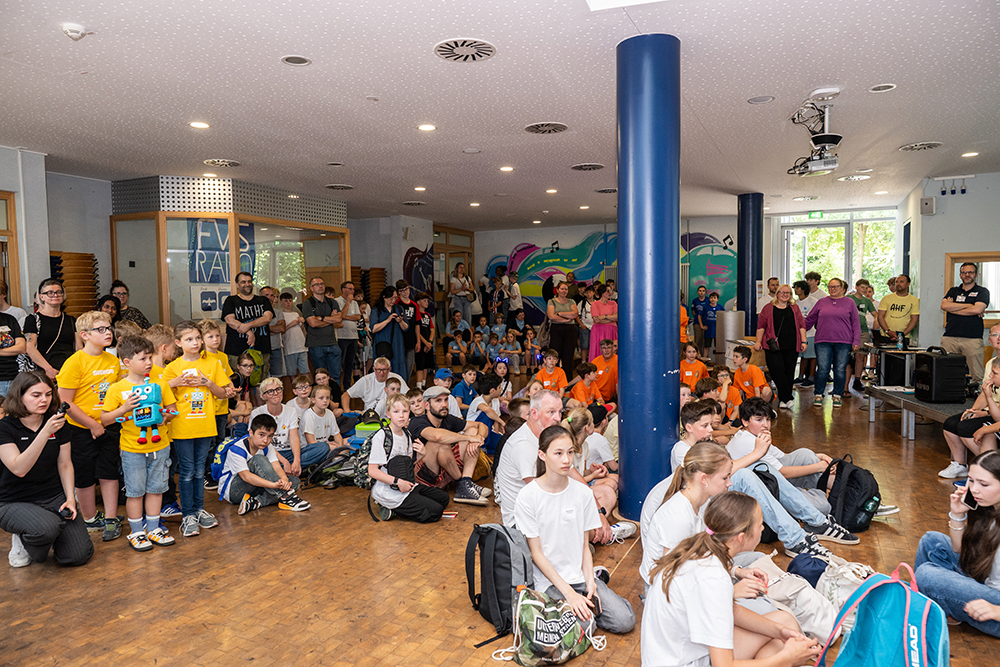 Kinder und Erwachsene befinden sich in einem Raum. Einige sitzen auf dem Boden, andere stehen, und alle hören einem Redner zu, der nicht zu sehen ist.