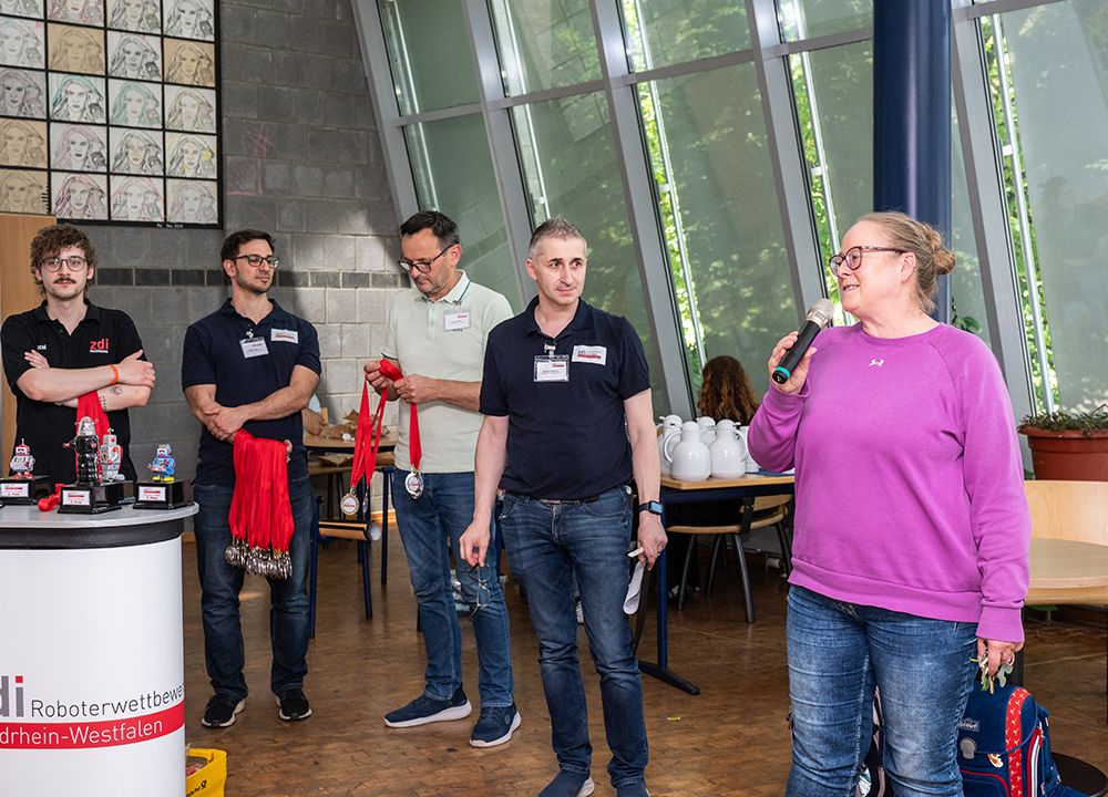 Fünf Erwachsene stehen bei einer Veranstaltung in einem Raum. Eine Frau spricht in ein Mikrofon, während die anderen zuhören. Auf einem Tisch stehen Auszeichnungen und Roboter.