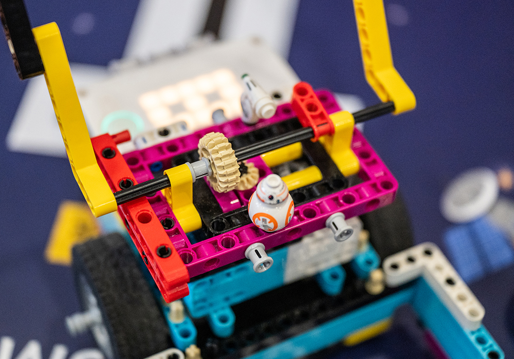 Ein bunter LEGO-Roboter mit Zahnrädern, Rädern und einer kleinen BB-8-Figur wird auf einer blauen Fläche in Nahaufnahme zusammengebaut.