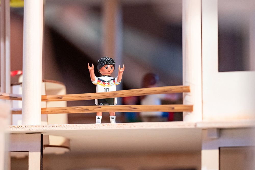 Eine Spielfigur im Playmobil-Stil steht mit erhobenen Armen auf einem hölzernen Balkon in einem Miniaturhaus.