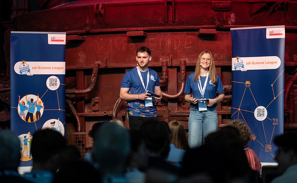 Zwei junge Moderatoren in blauen Hemden stehen bei einer Wissenschaftsveranstaltung auf der Bühne. Sie stehen zwischen zwei Bannern vor einem industriellen Hintergrund.