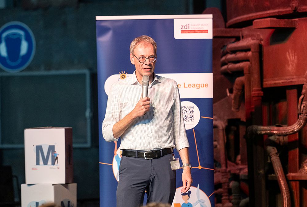 Ein Mann hält ein Mikrofon in der Hand und spricht auf der Bühne vor einem blauen Banner bei einer Veranstaltung in einer Halle.