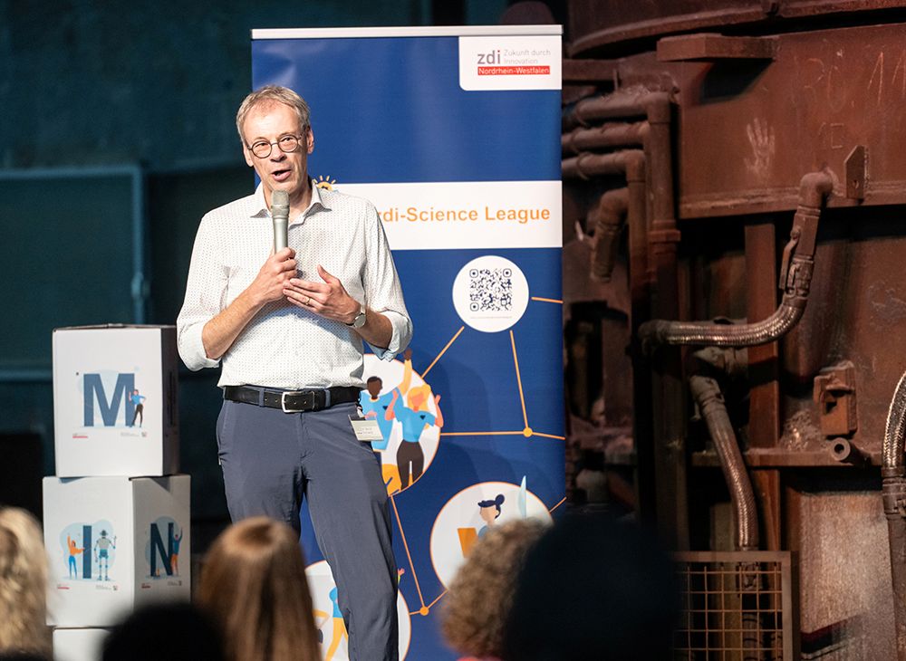 Ein Mann hält ein Mikrofon in der Hand und spricht auf der Bühne. Er steht vor einem Banner und boxt bei einer öffentlichen Veranstaltung, während das Publikum zusieht.